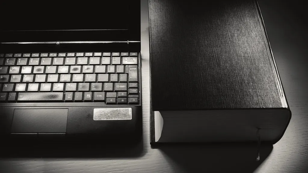 A laptop keyboard beside a closed hardback book, in black and white