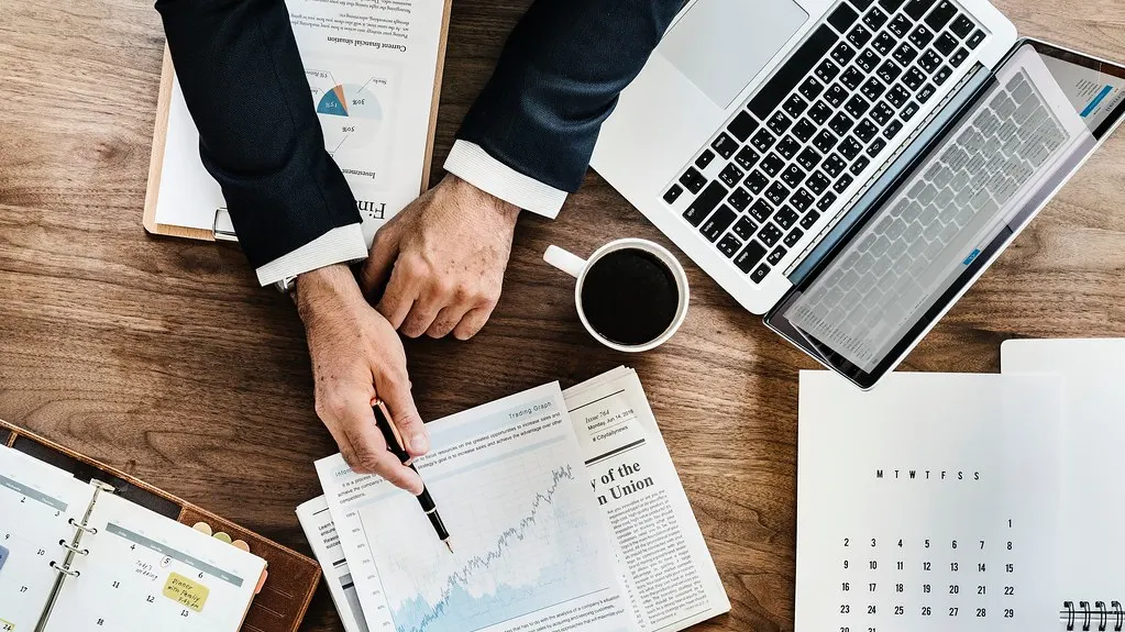 A person reviewing printed charts at a desk beside a laptop and a coffee